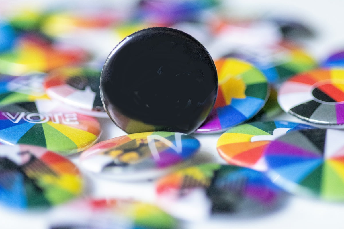 A solid black pin button resting on a pile of colorful rainbow circular badges